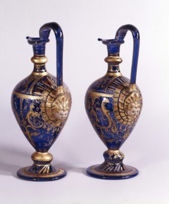 Zwei blaue Glasgefäße mit Bauch, schlankem Hals, Henkel und Ausguss. Verschiedene Ornamente in Gold bedecken den Objektkörper.