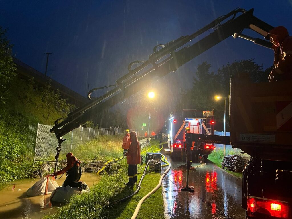 Drei Menschen platzieren bei Starkregen und im Dunkeln mit einem Kranfahrzeug schwere Säcke. 