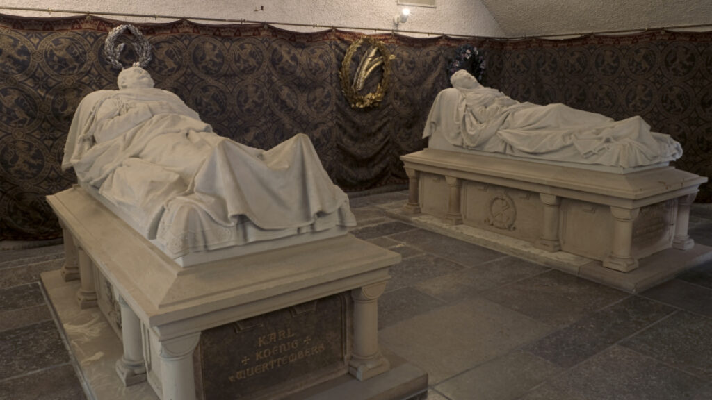 Zwei Steinsarkophage stehen vor einem Wandbehang in einem düsteren Raum.