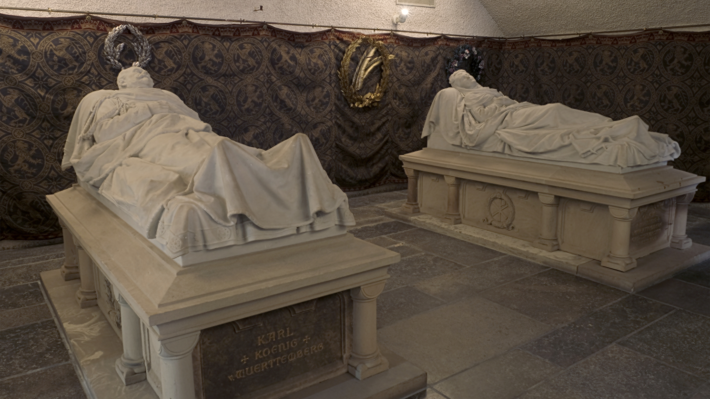 Zwei Steinsarkophage stehen vor einem Wandbehang in einem düsteren Raum.