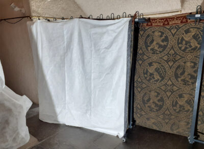 Wandbehang in der Mitte des Raumes auf Garderobenständer gehängt. Der Rückseitenschutz ist auf der linken Seite sichtbar.