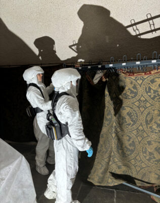 Zwei Personen in Ganzkörper-Schutz mit Gebläsemasken arbeiten am Wandbehang.