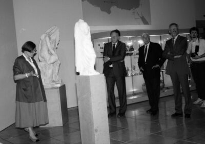 Schwarz-weiß-Fotografie, links im Bild eine Frau, rechts im Bild vier Männer. Dazwischen zwei Statuen auf Sockeln. 