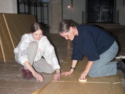 Foto von zwei Frauen, die auf dem Boden knien und mit Klebeband große Platten verkleben.