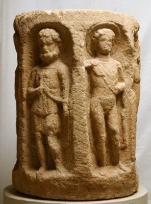 Runder, hellbrauner Stein mit zwei männlichen Figuren, die reliefartig vorne im Stein zu sehen sind.