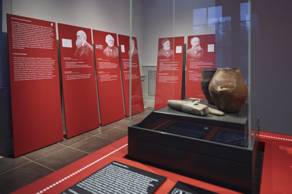 Blick in die Ausstellung: Im Vordergrund eine Vitrine mit Objekten, dahinter große Tafeln mit Portraits und Texten.