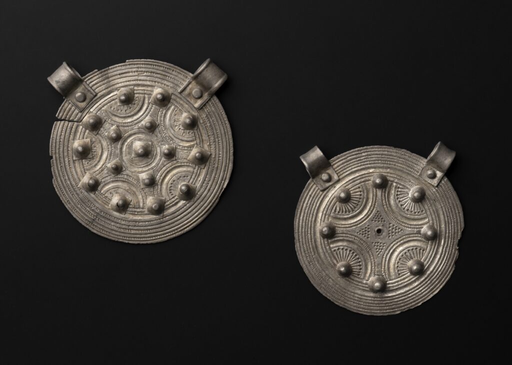 Zwei silberne Scheiben mit Ösen und Noppen vor dunklem Hintergrund.