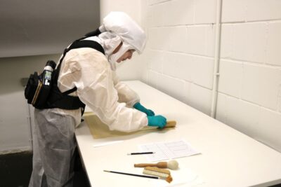 Eine Person in Schutzanzug reinigt Zeichnungen mit dem Pinsel.