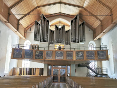 Eine moderne Kirche mit Orgelempore. Entlang der Brüstung befinden sich neun alte Gemälde.