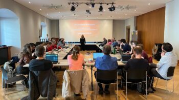 Raum, in dem mehrere Personen sitzen. Im Hintergrund läuft eine Präsentation.