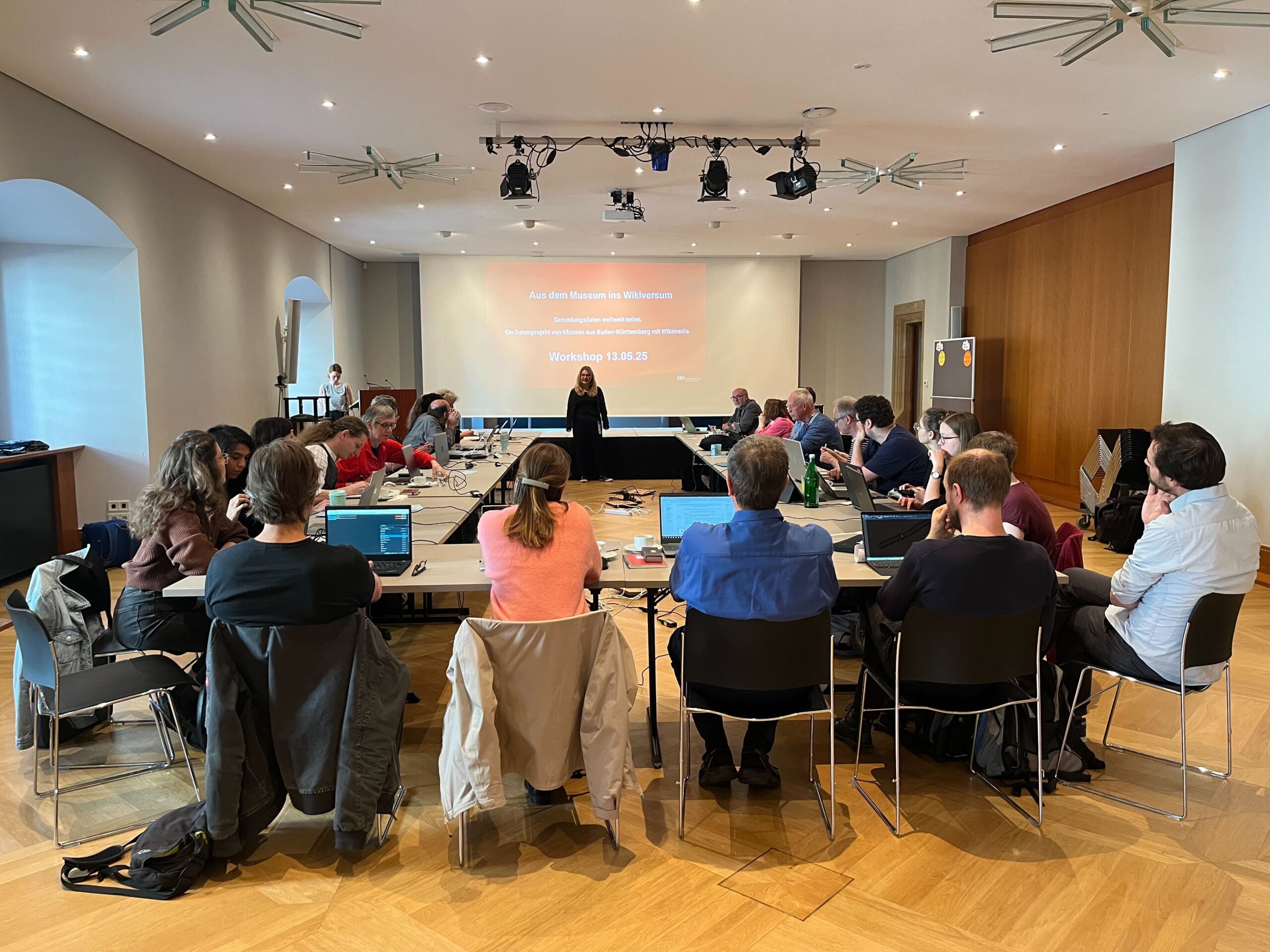 Raum, in dem mehrere Personen sitzen. Im Hintergrund läuft eine Präsentation.