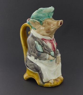 Figur eines Schweins. Das Schwein trägt eine Haube. Schürze und sitzt auf einem Stuhl.