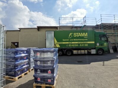 Großer, grüner LKW steht in einem Hof, davor stehen Kisten auf Paletten.