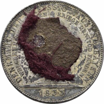 Runde Medaille mit umlaufender Aufschrift und roten Textilresten in der Mitte.