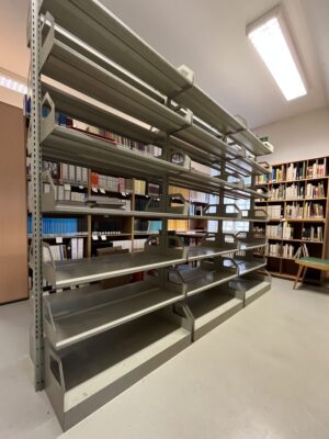 Leeres Bücherregal aus Metall. Im Hintergrund stehen Regale mit Büchern.