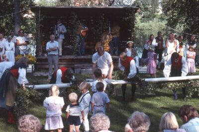 Verschiedene Personen bereiten Mittsommerbaum vor. Umgeben von Publikum. Im Hintergrund eine Bühne mit Musikern. 