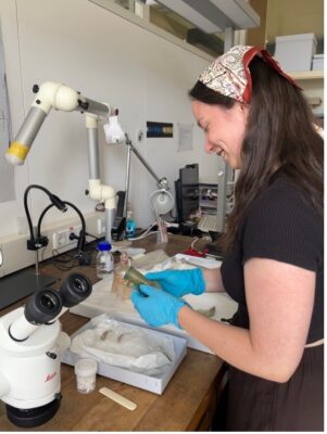 Junge Frau an einem Schreibtisch mit wissenschaftlichen Geräten, in der Hand hält sie ein antikes Glasgefäß.