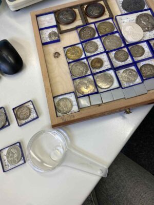 Verschiedene, einzeln verpackte Münzen und Medaillen und eine Lupe liegen auf einem Tisch.