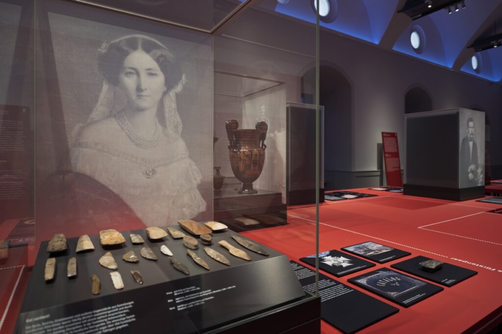 Im Zentrum steht eine Vitrine mit verschiedenen Steinobjekten, an der Rückwand ist das Portrait einer Frau zu sehen.