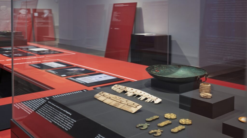 Blick in die Ausstellung. Im Zentrum eine Vitrine mit verschiedenen Objekten, wie Kämme und Zierelemente.