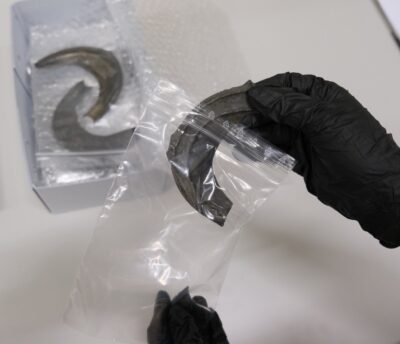 Ein Bronzeobjekt wird von einer Person, die schwarze Handschuhe trägt, in eine kleine Plastiktüte gesteckt. Im Hintergrund steht ein Karton, in dem weitere Objekte in Tütchen gelagert sind.