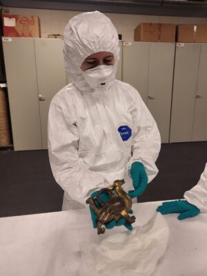 Junge Frau in weißer Schutzbekleidung im Depot mit dem Aquamanile in der Hand.
