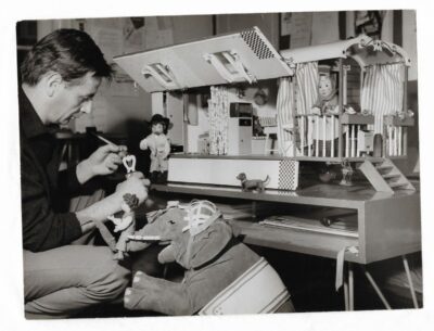 Schwarzweiß Fotografie. Ein Mann bastelt an einem Stuhl. Neben ihm steht ein Miniatur-Zirkuswohnwagen.