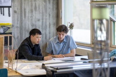 Farbfoto. Zwei junge Männer sitzen an einem Schreibtisch in einem Atelier. Sie blicken gemeinsam auf Papiere, einer der beiden zeigt mit einem Stift auf ein Papier.