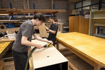 Farbfoto. Blick in eine Werkstatt. Zwei junge Männer arbeiten an der Holzummantelung der Vitrine. Einer bearbeitet das Holz mit einer Schleifmaschine, der andere hält ein Holzbrett in der Hand.