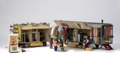 Ein buntes Modell eines Zirkusses vor einem hellen Hintergrund. In der Mitte stehen zwei Zirkuswägen. Links steht ein kleines Kassenhäuschen. Vor den Zirkuswägen stehen ein Stoffelefant und mehrere Stoffpuppen.
