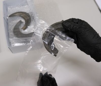 Zwei Hände in schwarzen Handschuhen legen ein Objekt aus dunkler Bronze in eine durchsichtige Plastiktüte. Im Hintergrund steht ein heller Pappkarton, in dem sich Luftpolsterfolie und weitere Bronzeobjekte in Tütchen befinden.